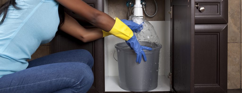 woman wearing rubber gloves with a bucket below a sink that's leaking water.